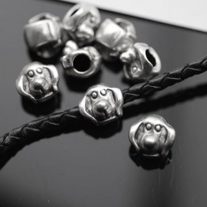 May include: Silver-toned dog-shaped beads with detailed facial features. The beads are scattered on a black surface and strung on a black leather cord. The beads are small, with a detailed dog face.