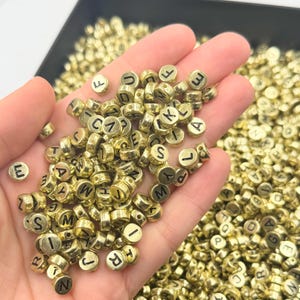 Könnte beinhalten: Ein großer Haufen goldfarbener Alphabetperlen mit schwarzer Beschriftung. Die Perlen sind rund und haben ein Loch in der Mitte zum Auf fädeln.