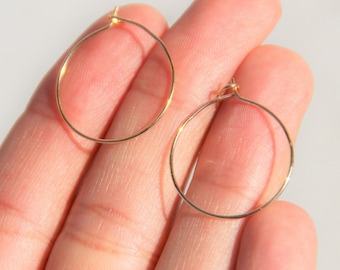 May include: Two delicate, gold-colored hoop earrings. The earrings are thin and circular, with a simple, minimalist design. The hoops are held in a hand against a soft, white background.