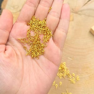 May include: A pile of gold-colored eyelet screws, also known as screw-back findings, in a hand. These are used for attaching jewelry, leather, and other crafts.