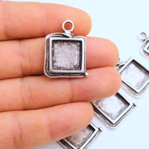 May include: Silver-toned metal pendant with a square frame and a textured surface. The frame has a slightly raised edge and a small loop at the top for attaching a chain.