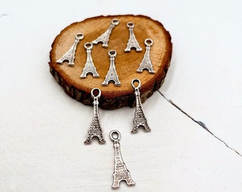 May include: A group of silver Eiffel Tower charms on a wooden surface. The charms are small and detailed, and they would be perfect for adding a touch of Parisian style to jewelry or crafts.