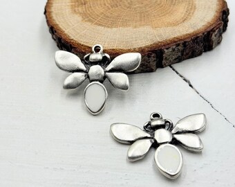 May include: Two silver-toned bee charms with white centers and loop attachments. The charms have detailed wings and body segments. The background includes a wooden slice and a white surface, suggesting a craft or jewelry theme.