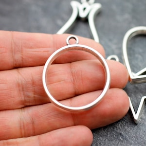 Könnte beinhalten: Silberfarbener Metallanhänger mit einem runden, offenen Rahmen. Der Anhänger wird in einer Hand gehalten.