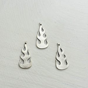 Könnte beinhalten: Drei silberfarbene Flammenanhänger. Jeder Anhänger hat eine Flammenform mit einem kleinen Loch oben zum Auffädeln. Die Anhänger liegen auf einer hellgrauen Oberfläche.