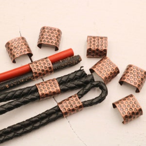 May include: A close-up of eight copper-colored metal cord ends with a textured, hammered finish. The cord ends are arranged on a white surface with a black leather cord.