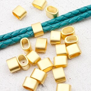 May include: A group of gold-coloured metal oval-shaped beads with a rectangular shape. The beads have a smooth, shiny finish and are scattered on a white surface. A teal braided cord is visible in the background.