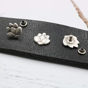 Könnte beinhalten: Nahaufnahme von vier silbernen Metallembellishments auf einer schwarzen Lederoberfläche. Die Verzierungen haben die Form einer Blume mit fünf Blütenblättern.