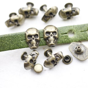 Könnte beinhalten: Antike messingfarbene Totenkopfnieten mit einem Stift und einer Unterlegscheibe. Die Nieten sind auf einem grünen Lederband angeordnet.