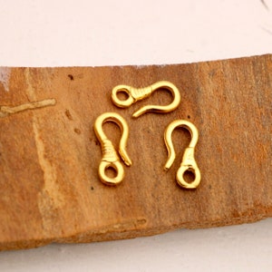 Peut inclure: Quatre fermoirs à ressort en métal doré, chacun avec une petite boucle en haut et un crochet en bas. Les fermoirs sont disposés sur une surface en bois.