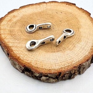 Könnte beinhalten: Drei silberfarbene Metall-Anhänger mit einem ovalen Loch in der Mitte. Die Anhänger liegen auf einer Holzoberfläche.