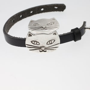 May include: A black leather bracelet with two silver cat head charms. The charms are detailed with eyes, whiskers, and a nose.