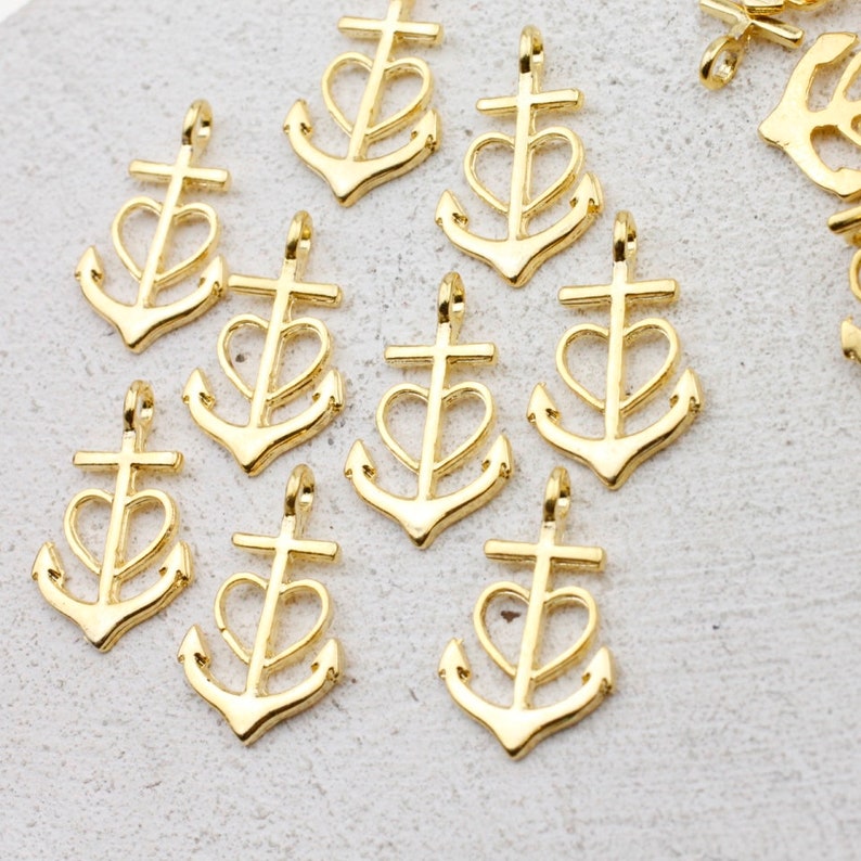 May include: A group of ten gold-colored metal anchor charms with a heart design. Each charm has a cross at the top and a heart shape in the center of the anchor.