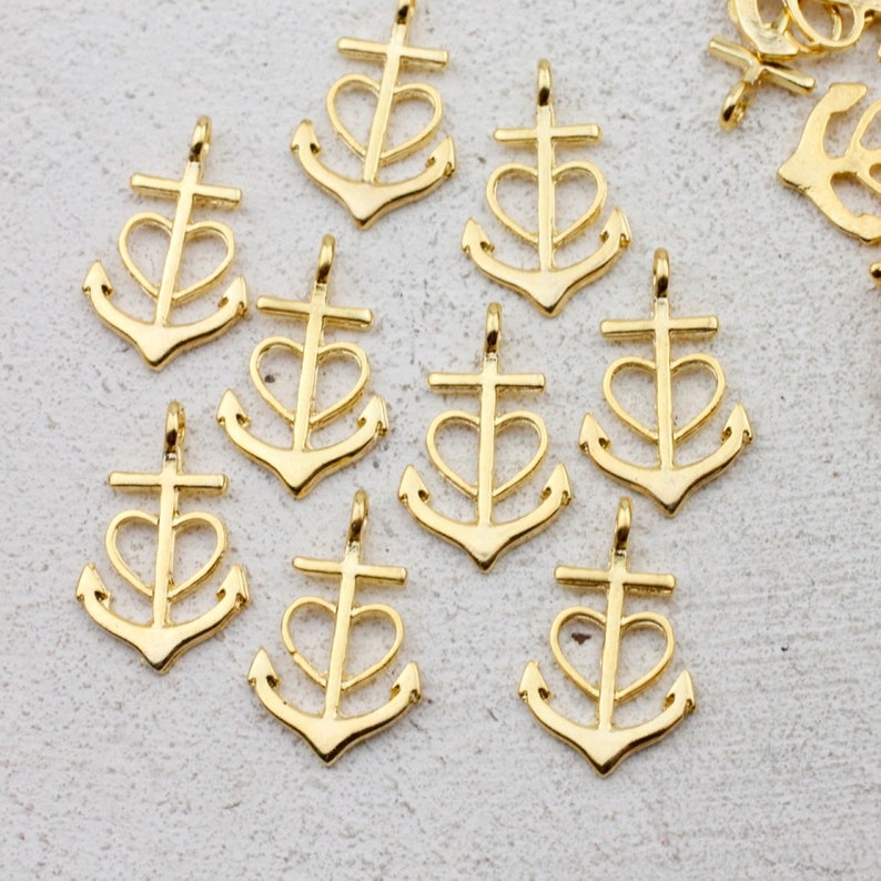 May include: A collection of ten gold-toned metal anchor charms with a heart design. The anchors are all the same size and shape, and they have a smooth, polished finish.