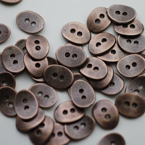 May include: A collection of oval-shaped, copper-toned buttons with two holes each. The buttons are scattered on a white surface.
