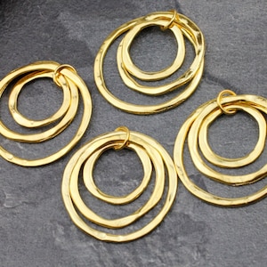 Peut inclure: Quatre anneaux en métal doré, chacun avec trois cercles concentriques, sont disposés sur une surface grise. Les anneaux ont une texture martelée et sont conçus pour être utilisés comme des trouvailles de bijoux.