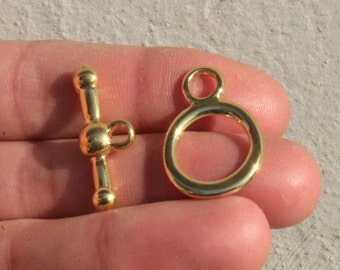 May include: Two gold-toned metal clasps for jewelry making. One clasp is a toggle clasp with a round ball on each end of the bar. The other clasp is a round ring with a smaller ring attached to the top.