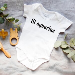 May include: White baby onesie with black text that reads "lil aquarius". The onesie is lying on a light gray fabric surface with a wooden baby toy, a wooden hair brush, and a pacifier.