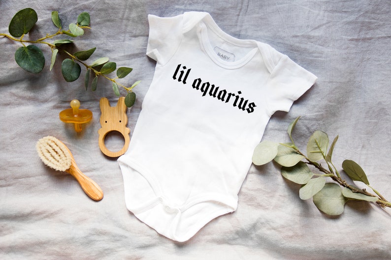 May include: White baby onesie with black text that reads "lil aquarius". The onesie is lying on a light gray fabric surface with a wooden baby toy, a wooden hair brush, and a pacifier.