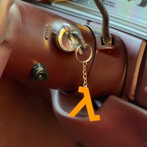 May include: An orange Lambda symbol keychain hanging from a car key in the ignition. The keychain is attached to a silver key ring and chain. The car's interior is a deep maroon color.