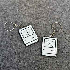 May include: Two white and black keychains shaped like vintage computers. One has a smiling face, the other a sad face. Each keychain has a silver keyring and chain, set against a gray fabric background.