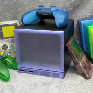 May include: A black cube-shaped console with a translucent purple front panel featuring USB ports, topped with a blue wireless controller. It is surrounded by a green translucent controller, a clear handheld game console, and a rainbow-colored cartridge rack.