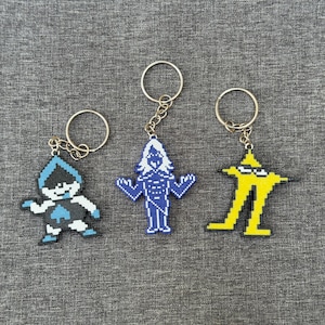 May include: Three pixel art keychains featuring characters from a video game. One keychain is blue and white, one is blue, and one is yellow. Each keychain has a silver keyring attached.