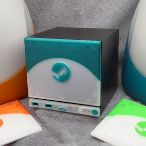 May include: A black cube-shaped computer case featuring a teal glitter front panel with a white semi-circle accent and a central Steam logo. The base includes three USB ports and a power button. Two additional faceplates in orange and green are included.