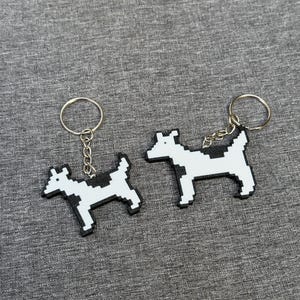 May include: Two pixelated dog keychains. Each keychain features a white dog with black spots and a black outline. The keychains are attached to silver key rings with a small chain.