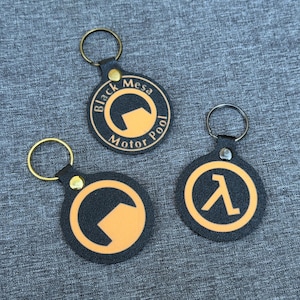 May include: Three black and orange keychains with gold-colored key rings. One keychain has the text "Black Mesa Motor Pool" around an orange circle. The other two keychains have orange symbols on a black background.