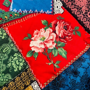May include: Close-up of a patchwork textile featuring various fabric squares. The squares display floral patterns, paisley designs, and lace trim in red, blue, green, and yellow. The central square is red with a floral bouquet.