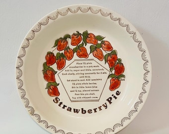 Op de afbeelding: Een ronde witte keramische taartschaal met een bruine geschulpte rand en een centrale illustratie van rode aardbeien in een cirkel. De tekst in het midden bevat een recept voor aardbeientaart en de woorden Strawberry Pie staan eronder.