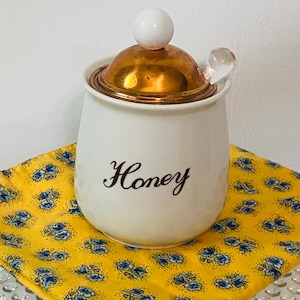 May include: A white ceramic honey pot with the word Honey printed in dark brown script. The pot features a metallic copper-colored lid topped with a small white spherical knob and a clear glass dipper handle extending from the side.