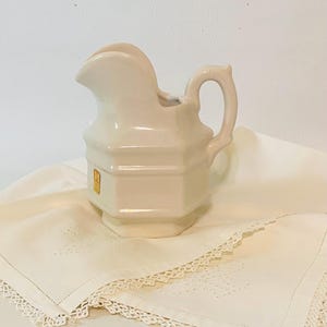 May include: A cream-colored ceramic pitcher with a hexagonal body, a curved handle, and a prominent pouring spout. The pitcher features a small, rectangular gold-toned label on its side and has a smooth, glossy finish throughout its structure.