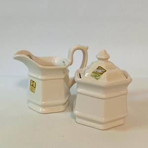 May include: A cream-colored ceramic creamer and a matching lidded sugar bowl with a square base and faceted sides. Both pieces feature a gold-toned rectangular sticker with the text "H" and "Crafted for a Century" on the front surface.