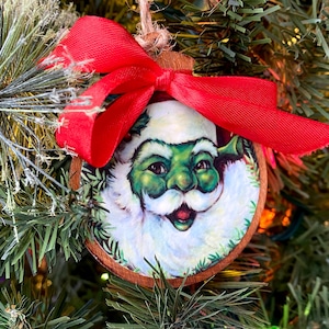 Pode incluir: Enfeite de Natal de madeira com um Papai Noel de pele verde com barba e chapéu brancos e uma fita vermelha. O enfeite é redondo e pendurado em um barbante natural. O enfeite está em uma árvore de Natal.