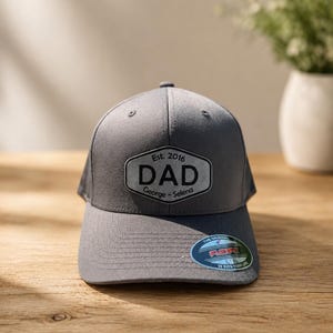 Puede incluir: Gorra de béisbol gris con visera curva. La gorra tiene un parche que dice "DAD" con "Est. 2016" arriba y "George - Selena" abajo. La gorra tiene una pegatina Flexfit.