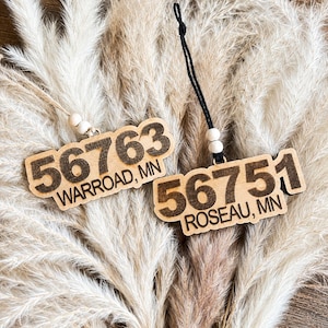 May include: Two wooden ornaments featuring laser-engraved text. One reads 56763 WARROAD, MN with a light-colored string and beads. The other reads 56751 ROSEAU, MN with a black string and beads. Both have a natural wood finish and dark lettering.
