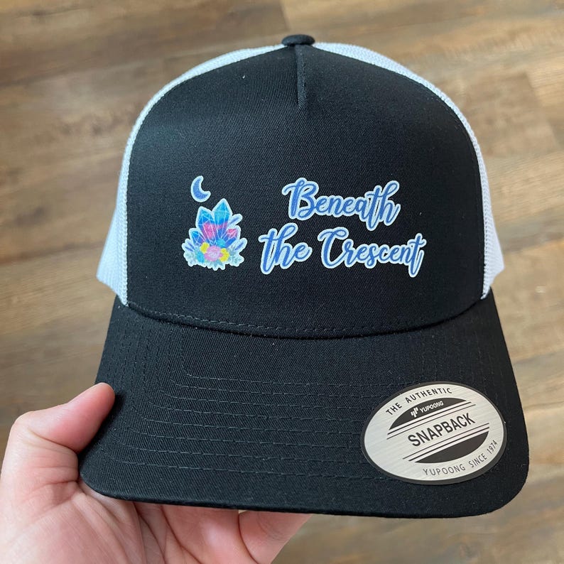 Puede incluir: Gorra de camionero negra y blanca con una parte trasera de malla negra. La parte delantera de la gorra presenta un dise&ntilde;o de cristal azul y morado con una luna creciente y el texto "Beneath the Crescent".