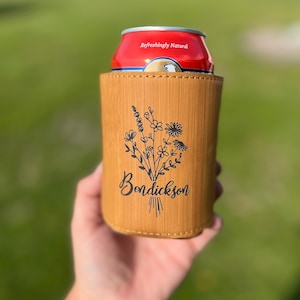 May include: A light brown bamboo can cooler with a floral design and the name "Bendickson" in black script. A red can with the text "Refreshingly Natural" is inside the cooler. The background is a blurred green.