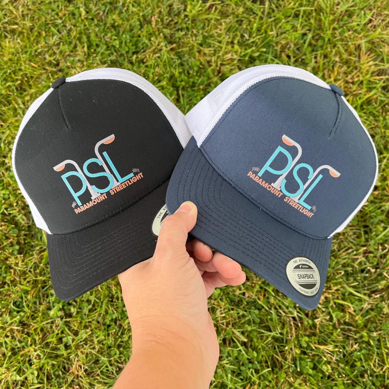 Puede incluir: Dos gorras de camionero, una negra y otra azul marino, con paneles de malla blanca. Ambas gorras presentan un logotipo blanco con el texto "PSL Paramount Streetlight" y un gr&aacute;fico de tres farolas.