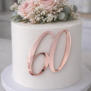 May include: A white frosted cake decorated with a rose gold number 60. The cake is topped with pink roses, small white flowers, and green foliage. The cake sits on a white cake stand.