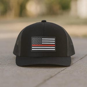 May include: Black trucker hat with a mesh back and a flat brim. The front features a rectangular patch with a stylized American flag design, including a red stripe. The hat is designed for casual wear.