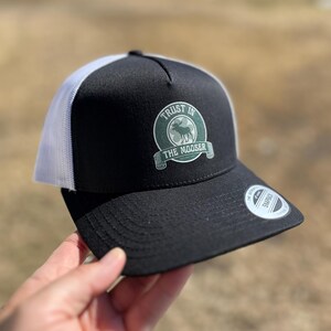 Puede incluir: Gorra de camionero negra y blanca con un logotipo circular verde que dice "Trust in the Mooser". El logotipo presenta una silueta de alce.