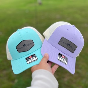 May include: Two trucker hats with a white mesh back and a solid front. The hat on the left is turquoise blue with a black leather patch that says "Campfire Therapy". The hat on the right is lavender with a black leather patch that says "Campfire Therapy". Both hats have a black "Richardson 115" label.