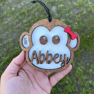 May include: A wooden monkey ornament with a white face, large eyes, and the name "Abbey" written in cursive. The ornament has a red bow and the year "2023" engraved on the side.
