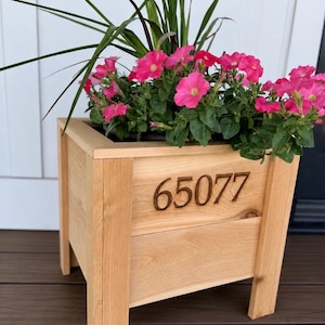 May include: A square light-colored wooden planter box with four legs, featuring the number 65077 engraved on the front panel. The planter is filled with vibrant pink petunia flowers and tall green grass blades extending from the top opening.