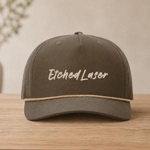 May include: A gray baseball cap with the text "Etched Laser" in a cursive font. The cap has a curved brim, a perforated back panel, and a tan rope detail above the brim. The cap is sitting on a wooden surface.