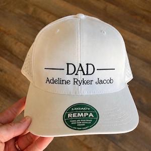 May include: White baseball cap with the word "DAD" embroidered in black, along with the names "Adeline Ryker Jacob." The cap has a curved brim and a mesh back. A green and white sticker on the brim reads "LEGACY REMPA."