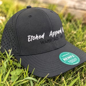 May include: A black baseball cap with a perforated side panel and a flat brim. The front features white embroidered text reading Etched Apparel and black text reading WARROAD, MN. A round green sticker on the brim reads LEGACY REMPA.